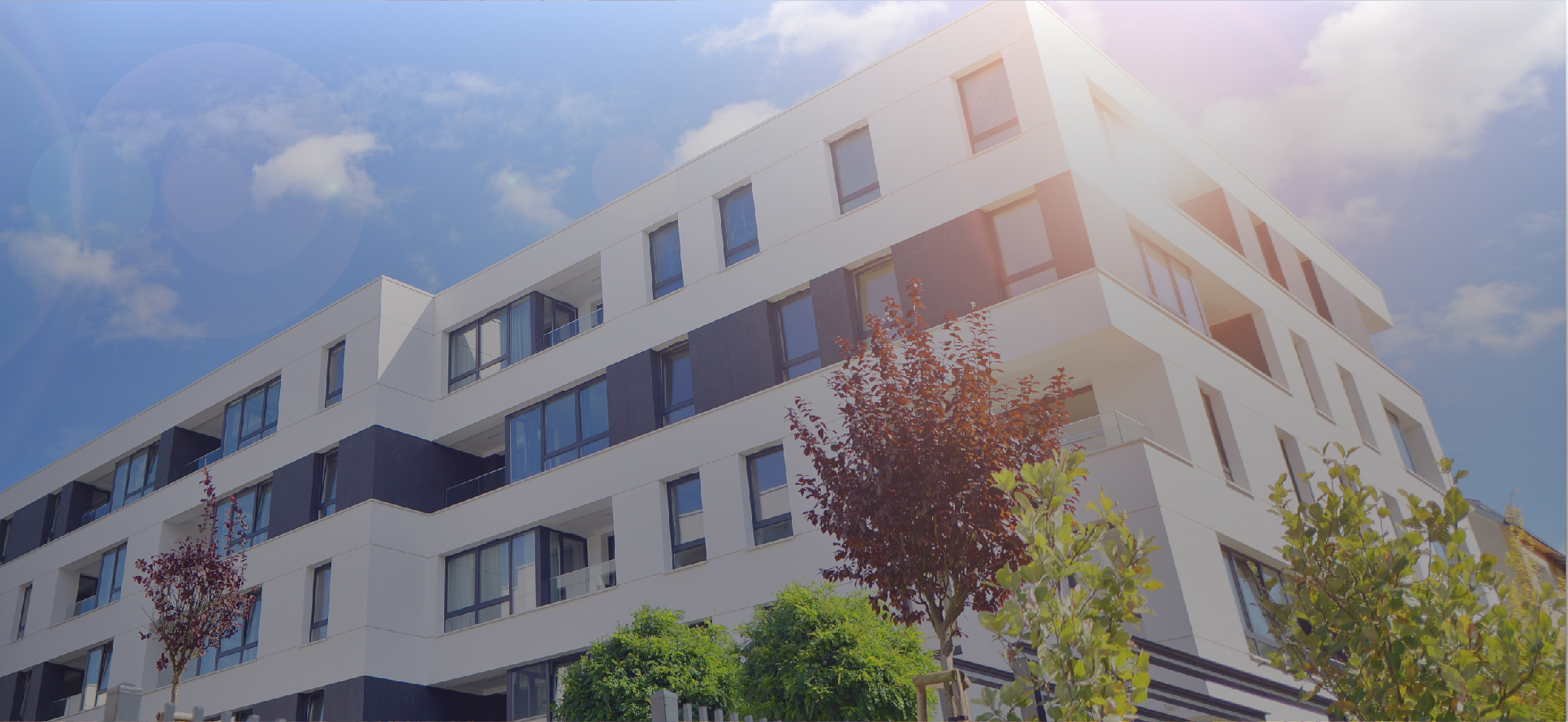 Modern white apartment building with curved balconies and landscaping under blue sky.